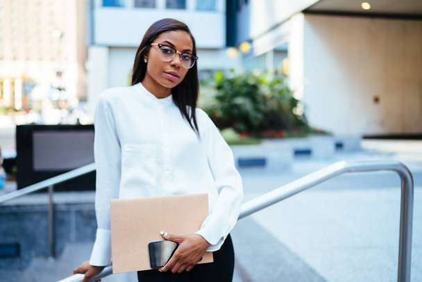 Half length portrait of confident female office employee with dark skinned looking at camera standing in urban setting.Serious african american businesswoman holding folder and smartphone in hands - Fotografie, Obrázek