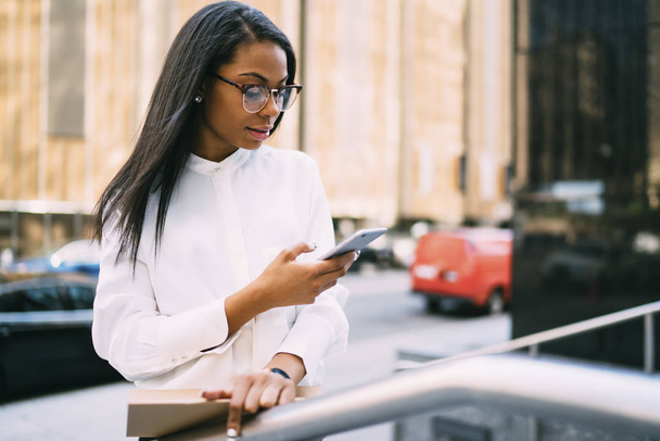 Pensive stylish young woman with dark skinned reading incoming sms on smartphone standing in downtown.African american office worker checking banking account online on cellular via 4G internet - Photo, image