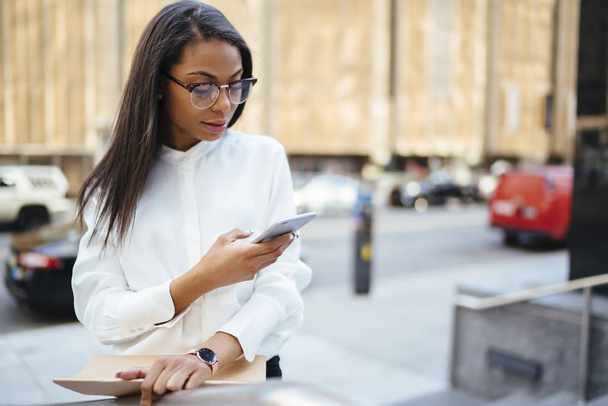 African american female employee dressed in stylish shirt sending notification via 4G internet on smartphone device standing in urban setting in downtown.Dark skinned office worker checking mail - Foto, Bild