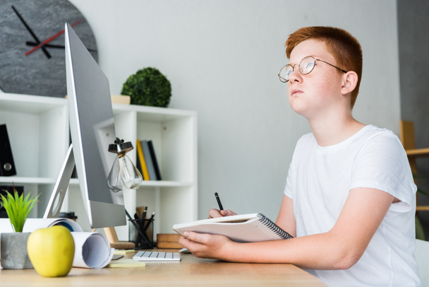 pensive preteen ginger hair boy holding pen with notebook at home and looking away - Fotografie, Obrázek