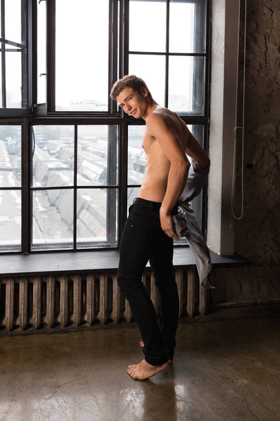 Shirtless handsome young man with brown hair standing near the large window iooking over his shoulder at the camera at a professional photostudio - Foto, Bild