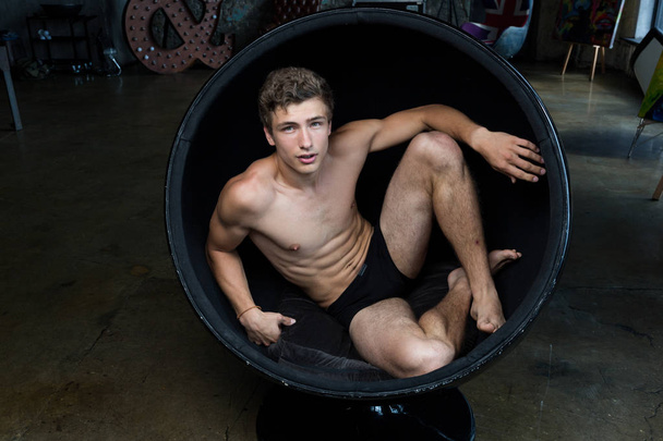 Handsome young man with brown hair in underwear sitting in a round black chair iat a trendy hipster interior of a professional photostudio - Photo, Image