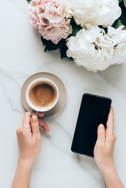 abgeschnittene Ansicht einer Frau mit Smartphone und Tasse Kaffee auf Marmoroberfläche mit Hortensienblüten  - Foto, Bild