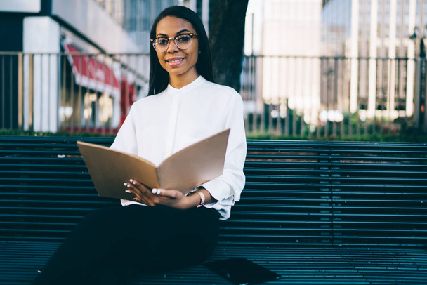 Portrait of positive african american female employee dressed in stylish white shirt smiling at camera while sitting outdoors in urban setting on bench and checking financial documents in folder - Photo, Image