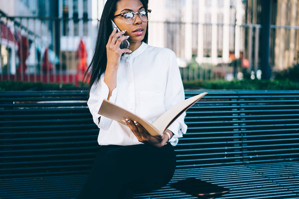 Serious dark skinned female office worker calling on smartphone while holding folder with documents in hands sitting outdoors on bench.Successful african american female employee talking on phone - Фото, зображення