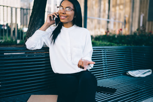 Cheerful african american young woman dressed in stylish white shirt discussing funny news during phone conversation on smartphone resting in city on bench.Positive female employee talking on phone - Фото, изображение