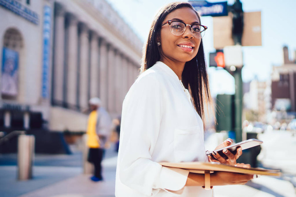 Portrait of successful dark skinned student of faculty of law holding smartphone and folder in hands smiling at camera standing on street against on university.Positive female employee with cellular - Fotoğraf, Görsel