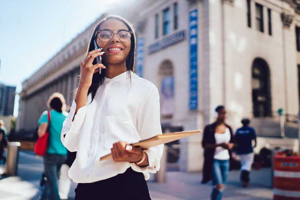 Portrait of african american successful student with folder in hands calling on smartphone and smiling at camera standing near university.Cheerful female office worker talking on telephone device - Foto, Bild