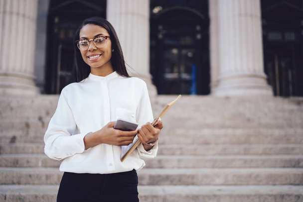 Half length portrait of prosperous african american student of faculty of law with folder and smartphone in hands smiling at camera standing against university building.Successful female office worker - Φωτογραφία, εικόνα