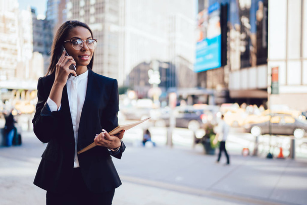 Successful african american female office worker dressed in stylish formal wear calling on smartphone standing in downtown.Prosperous employee with folder talking on cellular device in urban setting - Foto, immagini