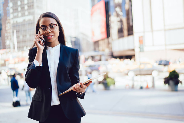 Połowę długości portret udanych african american kobieta interesu ubrany w czarny garnitur komunikacji na smartphone stojący w centrum miasta. Zamożnych kobiet partnerem w wizytowym wywołanie połączenia komórkowe - Zdjęcie, obraz