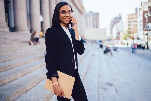 Portrait of successful african american businesswoman dressed in stylish formal wear talking with operator on cellular standing in downtown.Cheerful student lawyer with folder calling on smartphone - Photo, Image