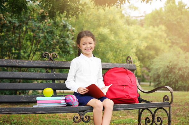 Cute little school child with stationery reading book on bench in park - Photo, image