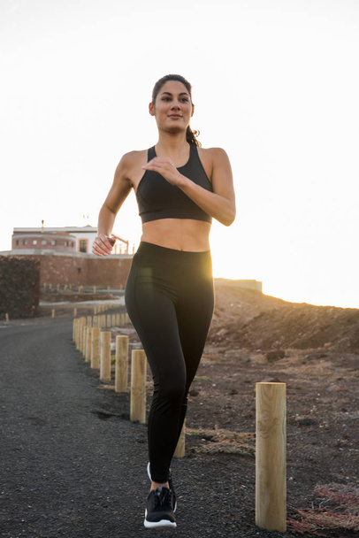 Woman wearing a black gym outfit is running on black tarred road next to a series of low poles in the sunset - Foto, Bild