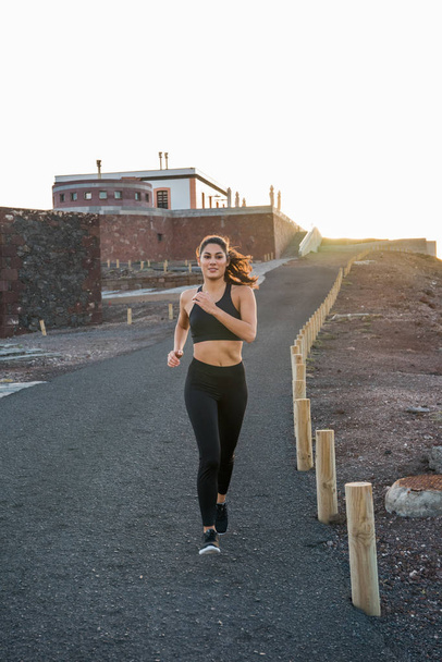 Young woman jogging on a road with a large building behind her in the sunset - Foto, Imagen