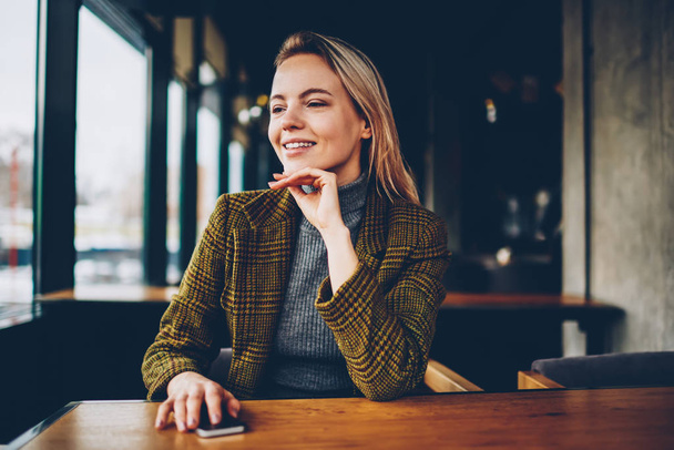 Happy blonde young woman dressed in stylish jacket looking spending leisure time in coffee shop.Cheerful hipster girl with smartphone in hand sitting at wooden table in cafe interior - Foto, Bild