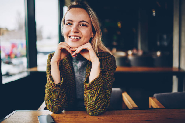 Half length portrait of happy female owner of stylish coffee shop resting at wooden table and laughing at camera in free time.Positive cute young entrepreneur smiling while sitting at desktop indoors - Φωτογραφία, εικόνα