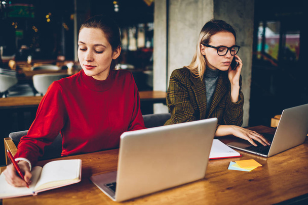 Two skilled graphic designers working remotely at modern netbooks connected to wireless internet in coworking.Blonde student calling on smartphone while colleague noting text in notepad from website - Foto, imagen