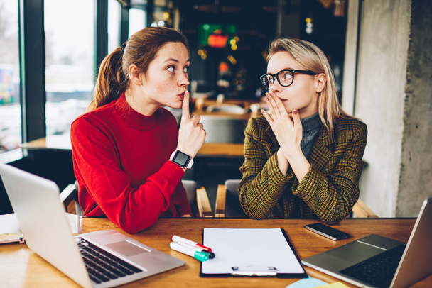 Brunette hipster blogger told secret to wondered colleague and showing sign shh during collaborating at laptop devices in coworking.Excited two best friends gossiping during studying break at netbooks - 写真・画像