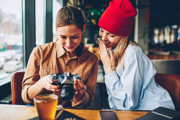 Cheerful female students watching made positive photos on display of vintage camera resting together in stylish cafeteria.Amazed hipster teenagers making editing of funny pictures during free time - Fotoğraf, Görsel