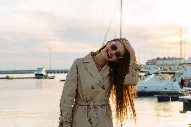 happy laughing young woman in beige coat posing against the sea and sunset background - Zdjęcie, obraz