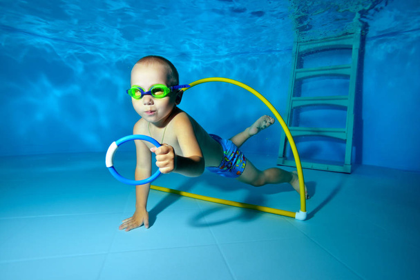 Malý chlapec plave pod vodou v bazénu a sbírá hračky ze dna. Portrét. Natáčení pod vodou. Horizontální orientace. - Fotografie, Obrázek