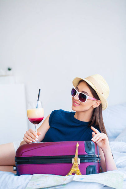 Vacation. Woman Who is Preparing for Rest. Young Beautiful Girl Sits on the Bed and Holds in the Hands Cocktail. Portrait of a Smiling Woman. Happy Girl Goes On Vacation. - Photo, image