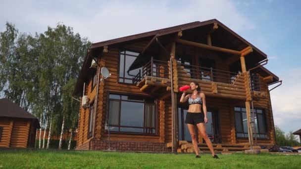 スポーティなブルネット女性黒トップの素敵な腹筋と木造の大邸宅の背景に汗で濡れて、トレーニングの後は、ボトルから水を飲む - 映像、動画