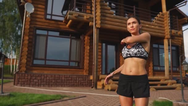 A slender and beautiful woman in sports clothes with an open press is preparing to start training on the lawn near her home stretching her arms and shoulders with warming up exercises - Footage, Video