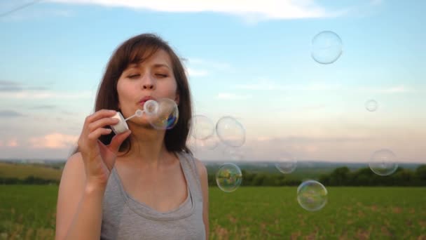 Krásná dívka s dlouhými vlasy foukat transparentní mýdlové bubliny proti modré obloze a usmívá se. Zpomalený pohyb. - Záběry, video