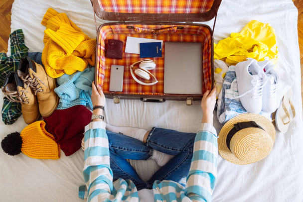 woman sit on bed with suitcase and clothes around. travel concept. decide warm or cold country to go. - Zdjęcie, obraz