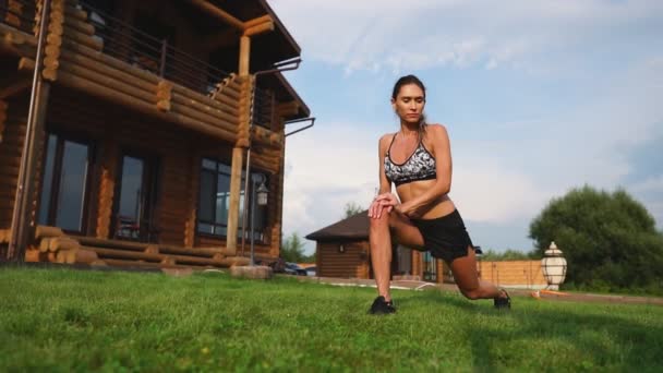 Une femme mince et belle en tenue de sport avec une presse ouverte se prépare à commencer l'entraînement sur la pelouse près de sa maison en fléchissant ses jambes, genoux, tibias et cuisses avec des exercices de réchauffement
 - Séquence, vidéo