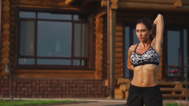 A slender and beautiful woman in sports clothes with an open press is preparing to start training on the lawn near her home stretching her arms and shoulders with warming up exercises - Footage, Video