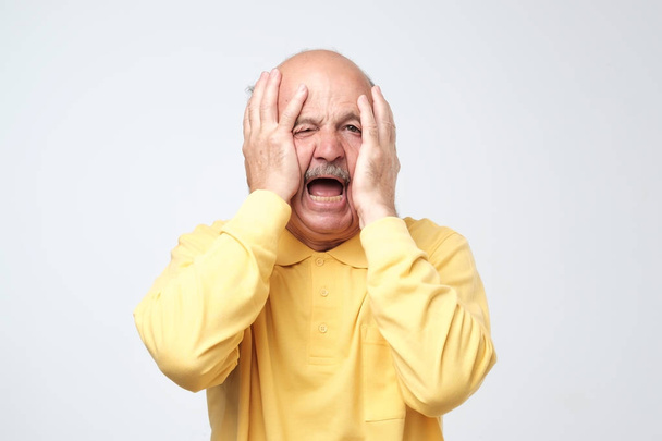 Homme hispanique senior en t-shirt jaune tirant la peau sur le visage avec les mains. Il est effrayé ou fatigué du stress
. - Photo, image