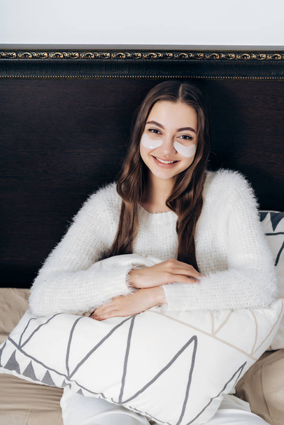 charming young woman in white pajamas lies in bed early in the morning, under the eyes white patches against swelling - Fotografie, Obrázek
