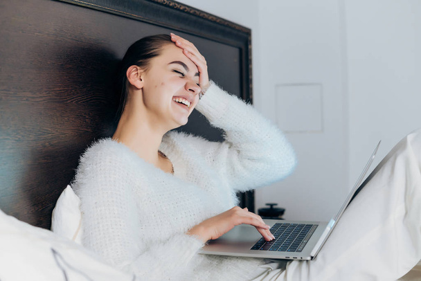 young girl in white pajamas sits in bed in the evening, looks funny video on her laptop - Foto, Imagem
