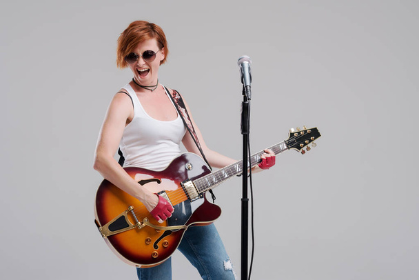 Portrait of a young woman musician with an electric guitar in her hands and studio microphone on a gray background. She wears black sunglasses and plays rock and roll loudly - Photo, image