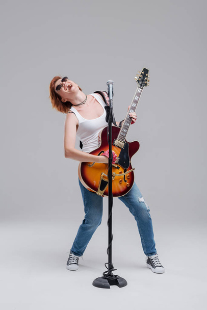 Portrait of a young woman musician with an electric guitar in her hands and studio microphone on a gray background. She wears black sunglasses and plays rock and roll loudly - Photo, Image