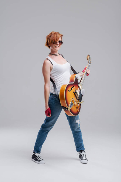 Young woman musician with an electric  guitar in hand on a gray background. He laughs and plays rock and roll loudly. Full-length portrait - Photo, image