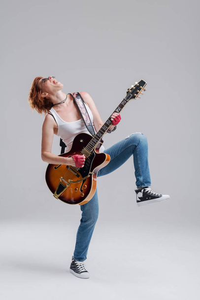 Młoda kobieta muzyk z gitara elektryczna w ręku na szarym tle. On śmieje się i głośno gra rock and rolla. Portretu pełnometrażowego - Zdjęcie, obraz