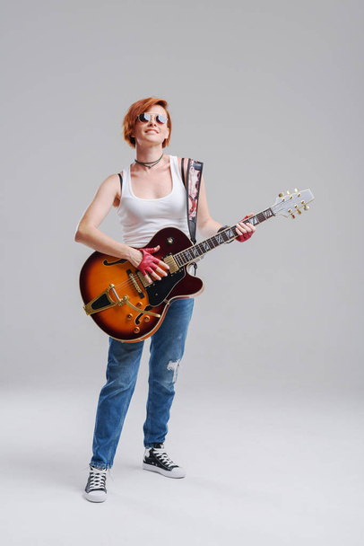 Young woman musician with an electric  guitar in hand on a gray background. He laughs and plays rock and roll loudly. Full-length portrait - Foto, imagen