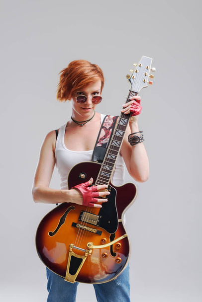 Portrait of a young woman with an electric guitar in her hands on a gray background. She wears black sunglasses, laughs  and Hugs a musical instrument - Foto, afbeelding