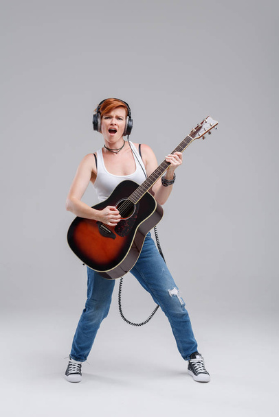 Young woman musician with an acoustic   guitar in hand and in headphones on a gray background. He laughs and plays rock and roll loudly. Full-length portrait - Photo, Image