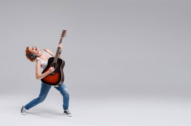 Young woman musician with an acoustic   guitar in hand and in headphones on a gray background. He laughs and plays rock and roll loudly. Full-length portrait. On the right there is space for text - Fotoğraf, Görsel