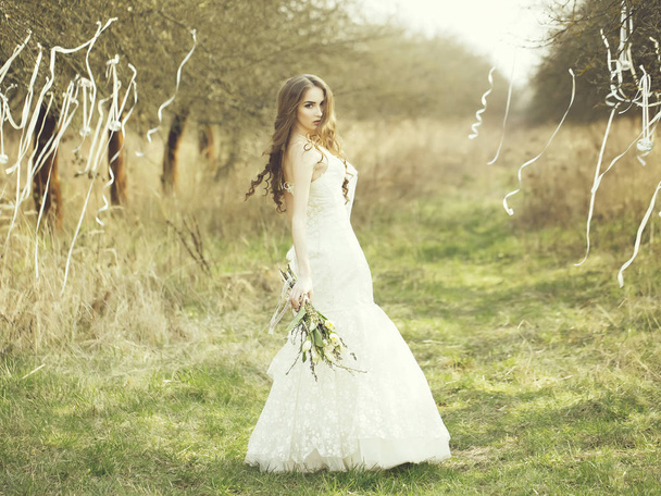 Pretty bride outdoor. Beautiful young bride in white wedding dress with flowers in field with ribbons on trees outdoor - Foto, Imagem