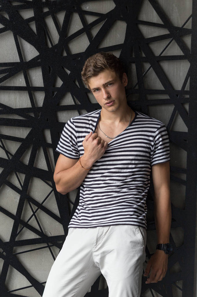 Handsome young male model in stripes t-shirt and white pants posing at modern interior photostudio - Фото, изображение