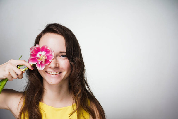 charming smiling girl in yellow dress is holding pink flower near face, posing - Photo, image