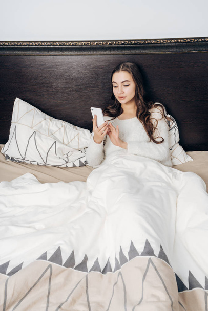 young tired girl is lying in bed late at night, resting, looking at her smartphone - Fotoğraf, Görsel