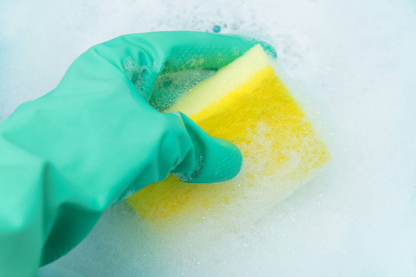 Une main dans un gant turquoise tient une éponge pour laver la vaisselle dans une mousse abondante. Gros plan
 - Photo, image