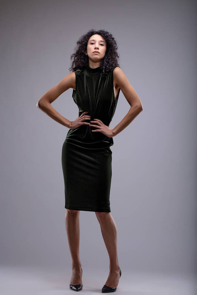 Λεπτός μοντέρνος αλαζονική νεαρή γυναίκα σε ένα μαύρο φόρεμα και τα ψηλά τακούνια που ποζάρει με τα χέρια στους γοφούς και μια σοβαρή έκφραση που απομονώνονται σε γκρι - Φωτογραφία, εικόνα